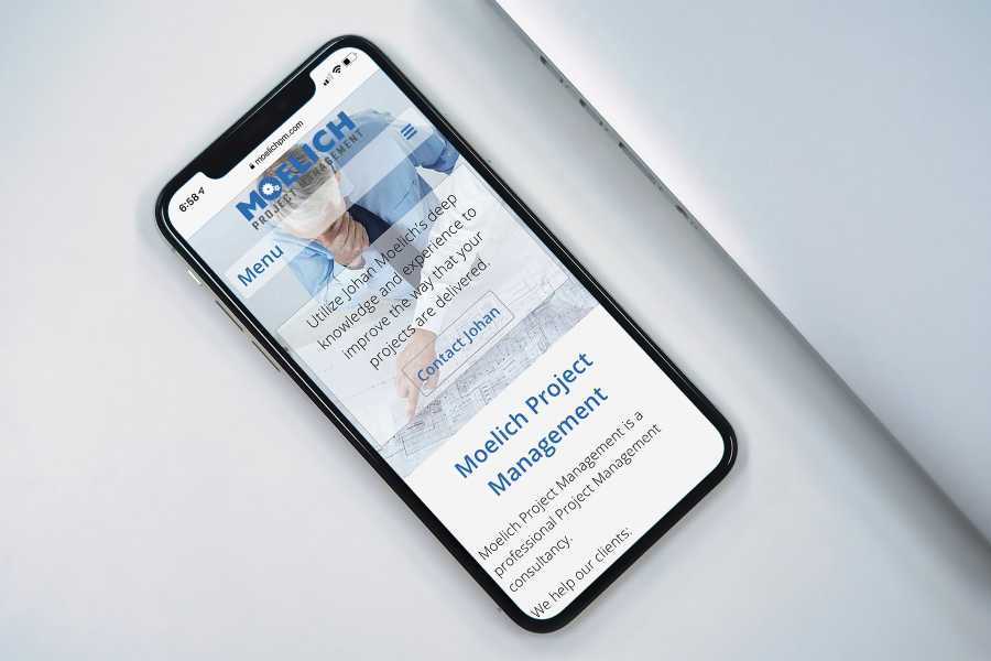 iPhone displaying Moelich Project Management home page, on table next to a laptop.