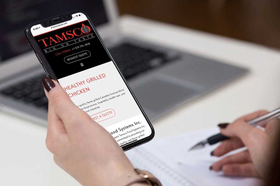 Tamsco Food Systems home page displayed on an iPhone being held by a woman.